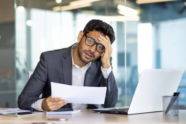 Üzgün ve hayal kırıklığına uğramış iş adamı yatırımcı kameraya bakıyor, elinde evrak raporları ve sözleşmeler tutan adam, banka başarısından memnun değil ve ofis içinde dizüstü bilgisayarla sonuçlanıyor..