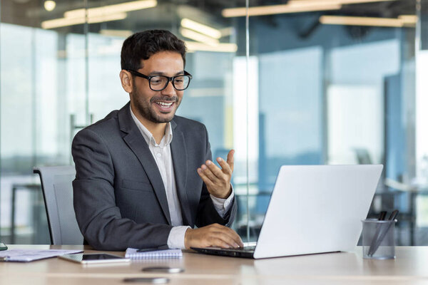 Young successful arab businessman inside office at workplace talking remotely with colleagues and partners, man using laptop for video call, satisfied boss smiling and gesturing with hands joyfully.