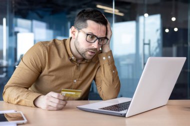 Ödeme ve kredi kartı işlemleri ile ilgili sorunlar. Genç bir adam ofiste dizüstü bilgisayar ekranının önünde oturuyor ve endişeyle kafasını tutuyor..