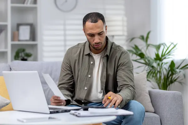 stock image Latin American man is focused working remotely at home on a laptop and with documents.