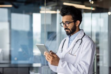 Genç bir Hintli doktor tablette çalışıyor. Hastane lobisinde steteskopu olan beyaz bir önlükle duruyor..