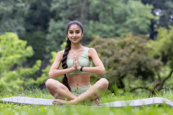 A portrait of a young Indian woman sitting in a park on a rug in a lotus pose and deals with yoga. Smile. He looks at the camera.