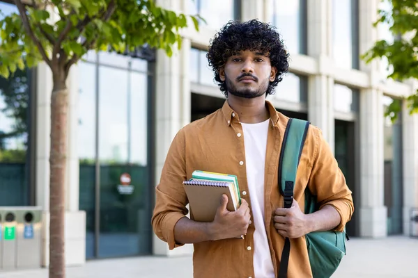 stock image Portrait of a young Indian student applicant standing outside the campus with a backpack and books in his hands and looking seriously into the camera.