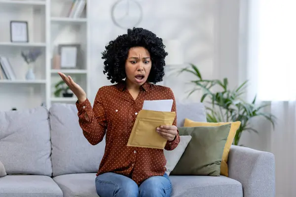 stock image Shocked African-American woman receiving bad news, documents from the bank, notice of debt. Sitting at home on the sofa and spreading his hands in frustration.