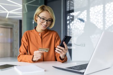 İş yerindeki ofisin içinde elinde telefon ve banka kartı olan genç ve güzel bir iş kadını masada oturan kadın çalışanlar online alışverişler ve para transferleri yapıyor..