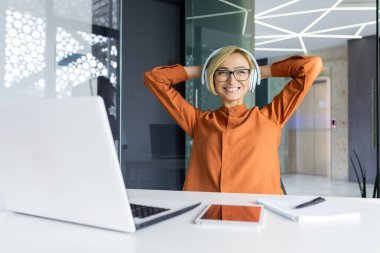 İş yerindeki başarılı ve memnun iş kadını bir iş projesini tamamladı. Dizüstü bilgisayarı ve kulaklığı olan kadın çalışanın elleri başının arkasında, gülümsüyor, müzik dinliyor..