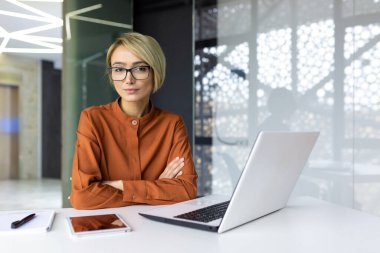 Konsantre düşüncenin portresi, ofis içi iş yerinde ciddi bir kadın, kollarını kavuşturmuş bir iş kadını dizüstü bilgisayarla masada oturuyor, finansör gömleği ve gözlüklü..
