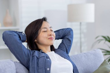 Young beautiful Asian woman relaxing at home close-up on sofa in living room, hands behind head with eyes closed dreaming and visualizing future results achievement and plans. clipart