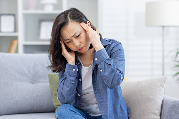 Sick woman alone at home, Asian depressed woman having severe headache, sitting on sofa in living room at home, holding hands to head from pain.
