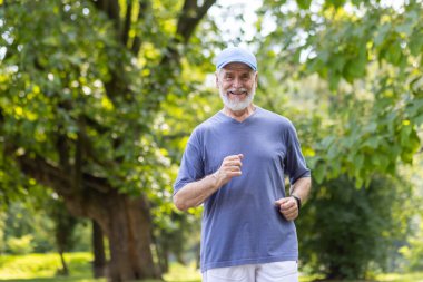 Yaşlı gri saçlı bir adam parkta koşuyor, emekli adam mutlu bir şekilde ağaçların arasında dinleniyor, spor kıyafetleri giyiyor ve kafasında bir şapka var..