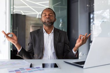 Olgun deneyimli Afro-Amerikan patronu ofis içinde meditasyon yapıyor masasında oturuyor, gözleri kapalı oturan adam geleceği görselleştiriyor zaferler ve başarılar, dizüstü bilgisayarlı iş adamı yatırımcı.