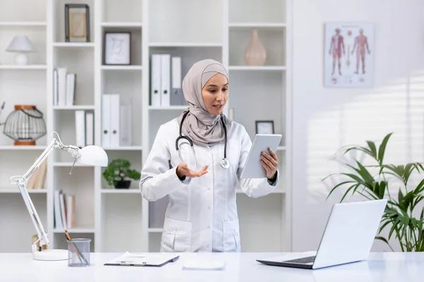 stock image Young experienced female doctor in hijab using tablet computer for video call, female clinic worker consults patients remotely online, smiling contentedly inside clinic office.