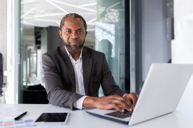 Ciddi düşünen ve kendinden emin bir iş adamının portresi, Afro-Amerikan patronu düşünüyor ve kameraya bakıyor, ofisin içinde takım elbiseli bir adam, yatırımcı ve dizüstü bilgisayarlı bir finansör.