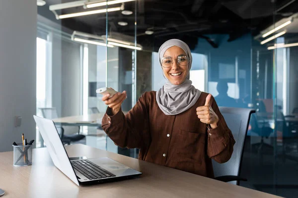 Portrait of a young Muslim woman in hijab working in the office, sitting at the desk and turning on the air conditioner with the remote control, pointing super finger and smiling at the camera.