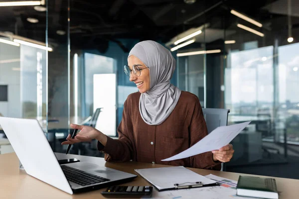 Young muslim businesswoman in hijab working in office with documents and talking on video call from laptop.