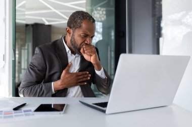 Hasta kıdemli Afro-Amerikalı patron iş yerinde öksürüyor, iş adamı laptopuyla masa başında oturuyor..