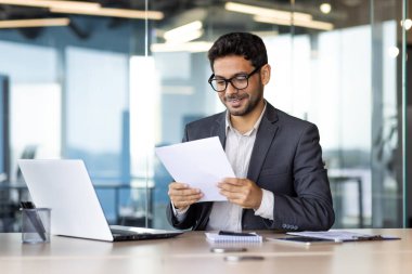 İş yerindeki deneyimli genç finansör şirket mali raporlarını kontrol ediyor ve okuyor, ofis içi evrak işlerinde çalışan iş adamı, başarı sonuçlarından memnun..