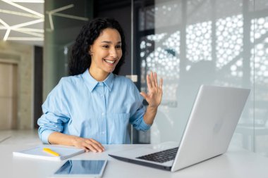 Ofiste laptopla çalışan genç başarılı İspanyol kadın, kameraya video kullanarak el sallıyor, iş kadını iş arkadaşlarıyla uzaktan konuşuyor..