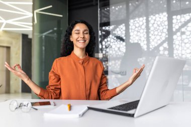 Genç ve başarılı bir kadın, Lotus pozisyonunda meditasyon yaparken ofis içinde oturuyor, gözleri kapalı İspanyol bir kadın, gülümsüyor ve gelecekteki planlarını ve başarılarını hayal ediyor..