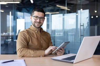 Ofiste çalışan gülümseyen genç bir adamın portresi, masada oturuyor ve elinde bir tablet tutuyor, kameraya gülümsüyor..