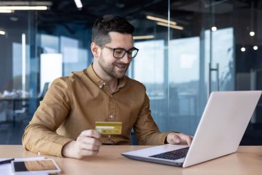 Genç bir iş adamı internette dizüstü bilgisayarla ve kredi kartı ekstreleriyle çalışıyor. Masada oturmuş gülümsüyor..