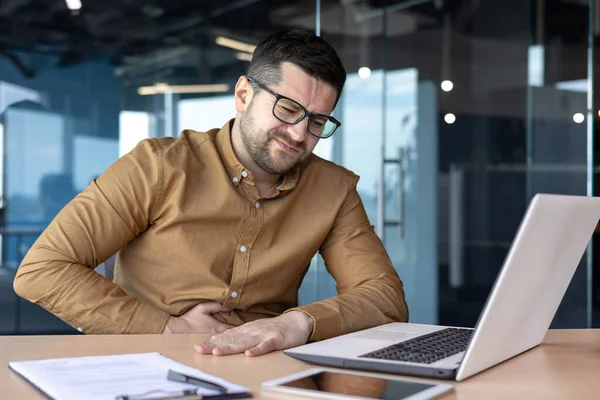 stock image Unwell at work. Young male office worker, businessman sitting in the office and holding his stomach, grimaced in pain and discomfort.