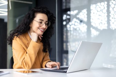 Mutlu ve gülümseyen İspanyol iş kadını dizüstü bilgisayarda yazı yazıyor. Kıvırcık saçlı ve gözlüklü ofis çalışanı ofis içinde başarı elde etmekten memnun.. .