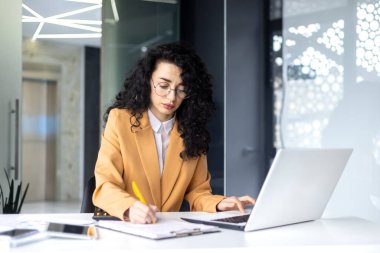 Ofiste çalışan ciddi ve düşünceli bir iş kadını, kadın finansör sözleşmeleri ve grafikleri düşünüyor, İspanyol başarılı kadın iş yerinde dizüstü bilgisayar kullanıyor.