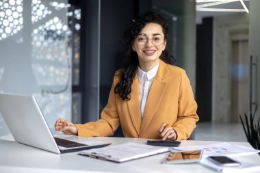 İş yerindeki ofis içinde, gülümseyen ve kameraya bakan, mali rakamları hesaplayan ve raporları hesap makinesiyle kontrol eden olgun, başarılı kadın finansör portresi..