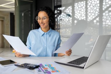 Genç başarılı finansör evrak işleriyle tatmin olmuş, İspanyol kadın raporları ve sözleşmeleri gözden geçiriyor, iş yerindeki bilgisayarla çalışıyor, iş kadını patronu başarılardan memnun..