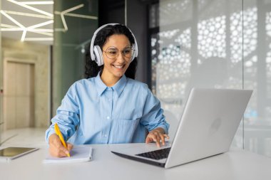 İş kadını ofis içi internet üzerinden video izleme kursu, kulaklıklı İspanyol kadın iş yerindeki verileri kaydederek mesleki gelişim ve mesafenin sonuçlarından memnun kalmış bir şekilde gülümsüyor.