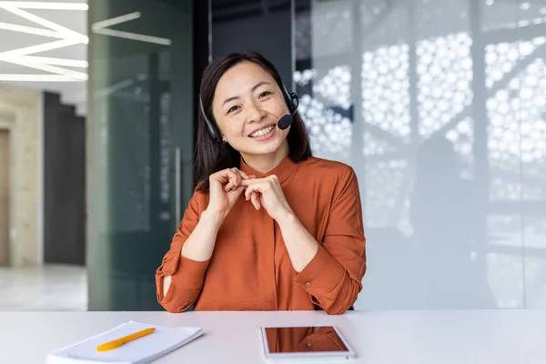Webcam view, online video call remote meeting, businesswoman with headset phone smiling and looking at camera, asian woman talking with colleagues using laptop while sitting inside office.