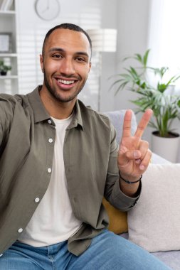 Dikey fotoğraf. Kameraya eli ve iki parmağıyla barış işareti gösteren gülümseyen Afro-Amerikan bir adamın yakın plan fotoğrafı..