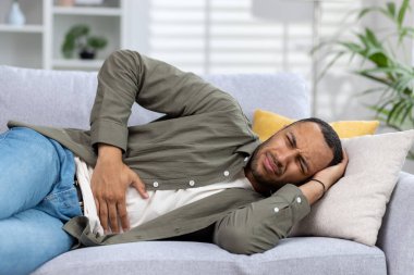 Genç Afro-Amerikalı adam evinde kanepede uzanmış yüzünü buruşturmuş ve acı içinde elleriyle karnını tutuyor. Yakın plan fotoğraf..