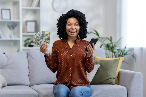 Portrait of a happy young African American woman holding a phone and a fan of money in her hands and showing the camera. Sitting at home on the couch and smiling at the camera.