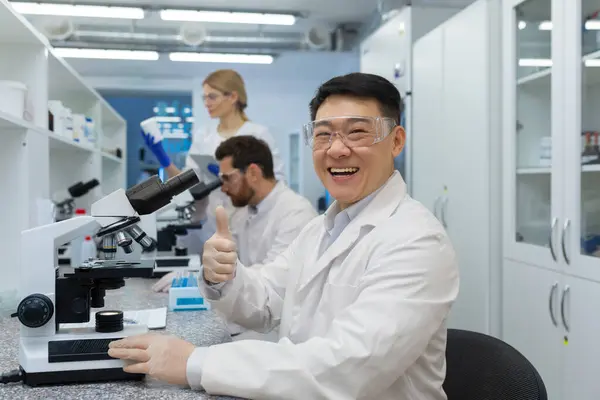 Portrait of a smiling and a young Asian male doctor, scientist sitting behind a microscope and pointing at the camera with super finger, colleagues are working in the laboratory behind.