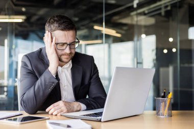 İşle ilgili sorunlar. Üzgün genç bir ofis çalışanı, iş adamı bir dizüstü bilgisayarın önünde oturuyor ve düşünceli bir şekilde başını eline yaslıyor..