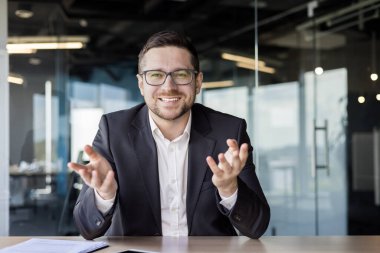 Genç bir erkek öğretmenin portresi çevrimiçi olarak ofisten video yoluyla öğretmenlik yapıyor. Kameranın önünde oturuyor ve gülümseyerek elleriyle el kol hareketi yapıyor..