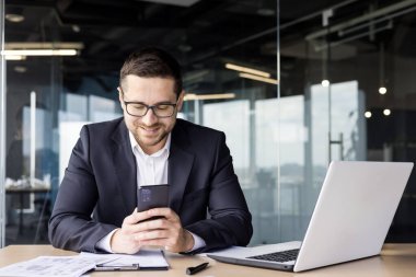 Genç bir erkek ofis çalışanı bir masaya oturur ve telefonu kullanır, gülümseyerek mesajlar yazar, haberleri okur, online oyunlar oynar..