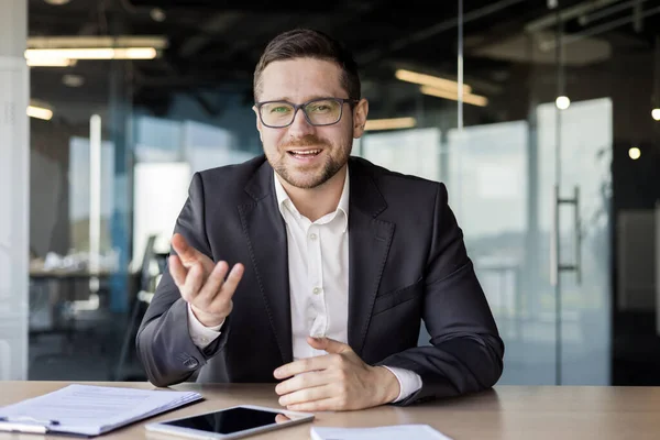 Portrait of a young businessman, a business coach conducts training and online training, professional development. Sitting in the office at the table in front of the camera, gesturing with his hands.