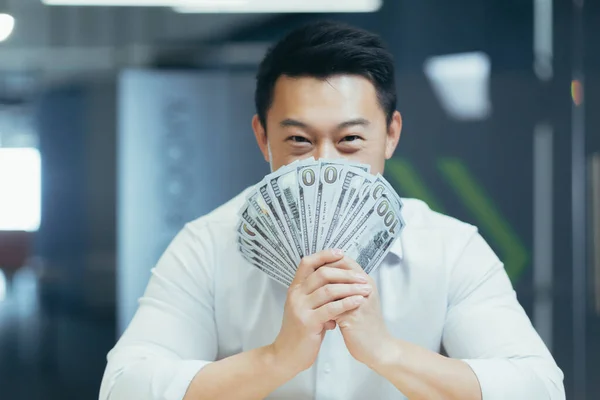 白いシャツで笑顔の若いアジア人男性の写真を閉じ カメラの前にオフィスに座って 彼の顔の前に現金とドルのファンを持っている — ストック写真
