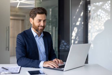 İş elbisesi giymiş olgun bir iş adamı iş yerinde dizüstü bilgisayarda çalışıyor. Başarı sonuçlarından memnun olan adam gülümsüyor, klavyede daktilo kullanıyor..