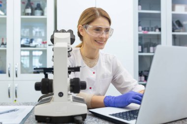 Gülümseyen genç bir kadın bir laptopun laboratuarında çalışıyor. Beyaz önlük ve koruyucu gözlük takmış, masasında oturuyor..
