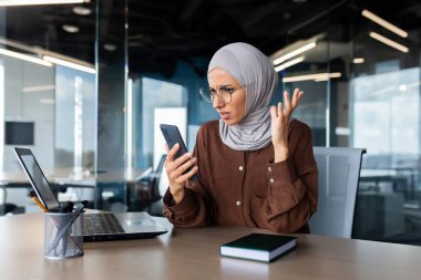 Endişeli genç Müslüman iş kadını ofiste çalışıyor ve şok olmuş bir şekilde telefon ekranına bakıyor..