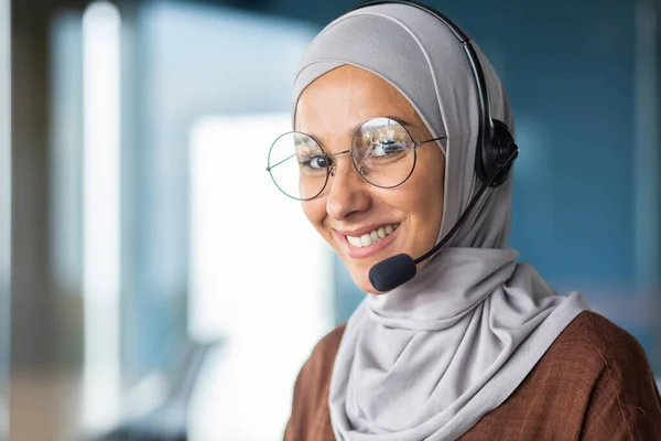 Close-up photo of a young Muslim woman in a hijab wearing a headset working in the office and smiling at the camera.