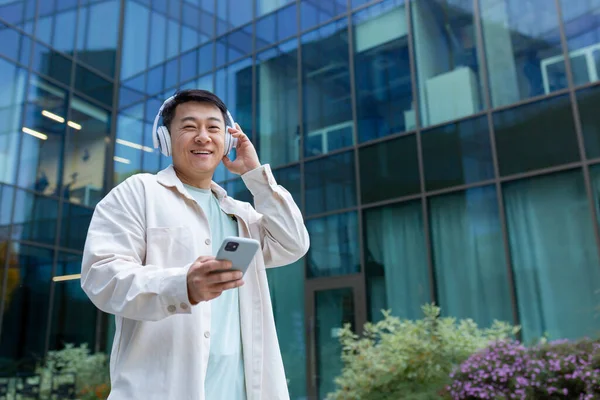 Portrait of asian man outdoors with phone and headphones for listening to music, happy man using smartphone app smiling and looking at camera.