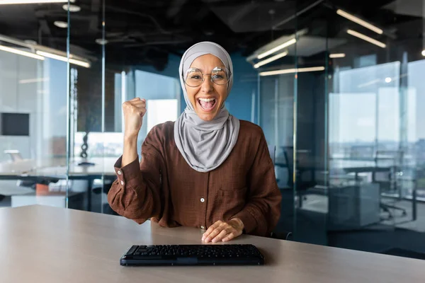 Happy young muslim woman in hijab is happy with success and achievement in front of camera, sitting at office desk and showing victory gesture with hand.