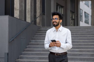 Genç bir adam, bir işadamı, bir programcı şehirde yürüyor, telefonu elinde tutuyor, mutlu bir şekilde gülümsüyor, mesajlar yazıyor ve internette geziniyor..