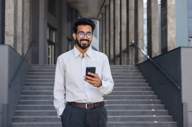 Başarılı genç Hindu adam ofis binasının dışında yürüyor, mühendis yazılım geliştiricisi gülümsüyor ve mutlu telefonda test uygulaması kullanıyor, sonuçtan memnun..