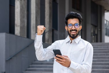 Arap online kazanan portresi, iş gömlekli bir adam ofis binasının dışında dolaşıyor, iş adamı gülümsüyor ve kameraya bakıyor, zafer jestini kaldırıyor, telefondaki uygulamayı kullanıyor..